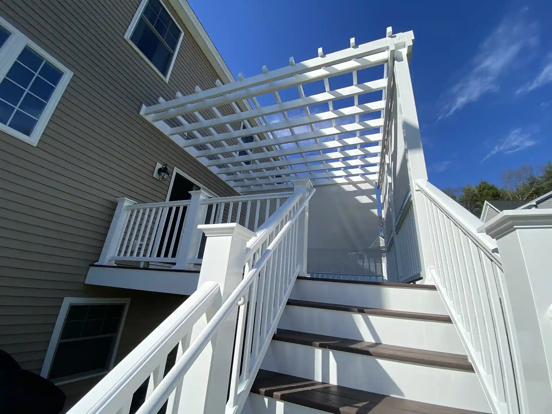 Photo of a raised deck with stairs and a white pergola casting shadows under a blue sky.
